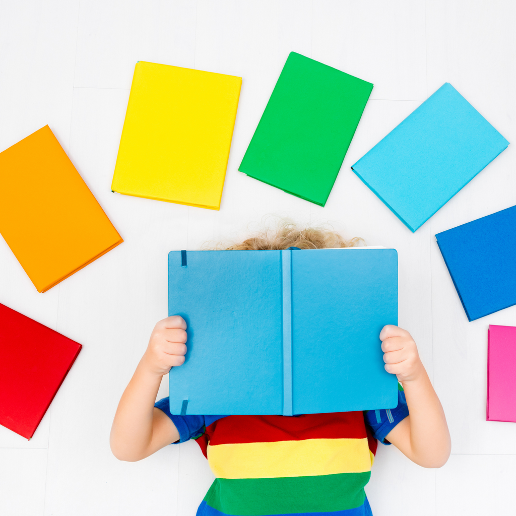 child lying on back reading a blue books and surrounded by colourful books on white background.