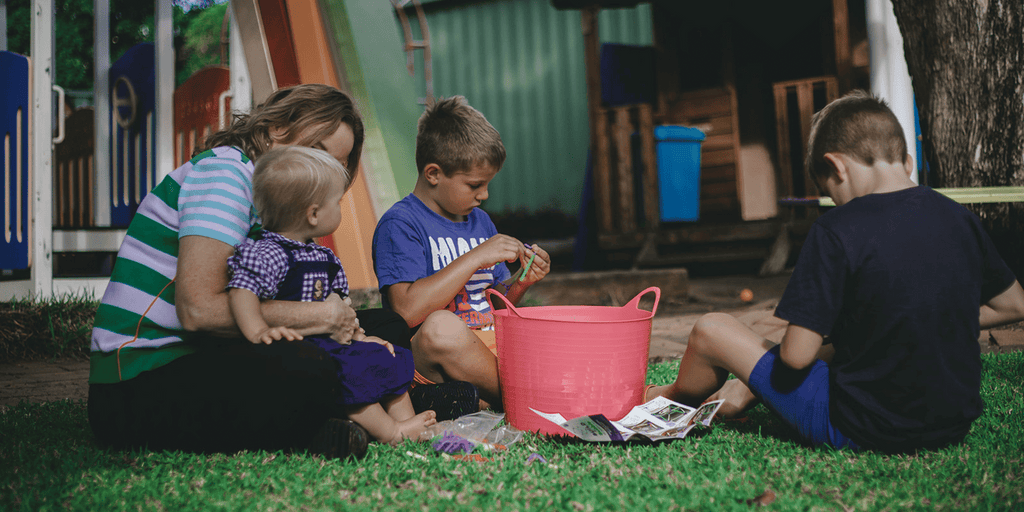 Image shows Kylie Toynton and three children playing outside on the grass. They are sitting around a red bucket and playing with the South Beach Bubbles Build a Bubble kit